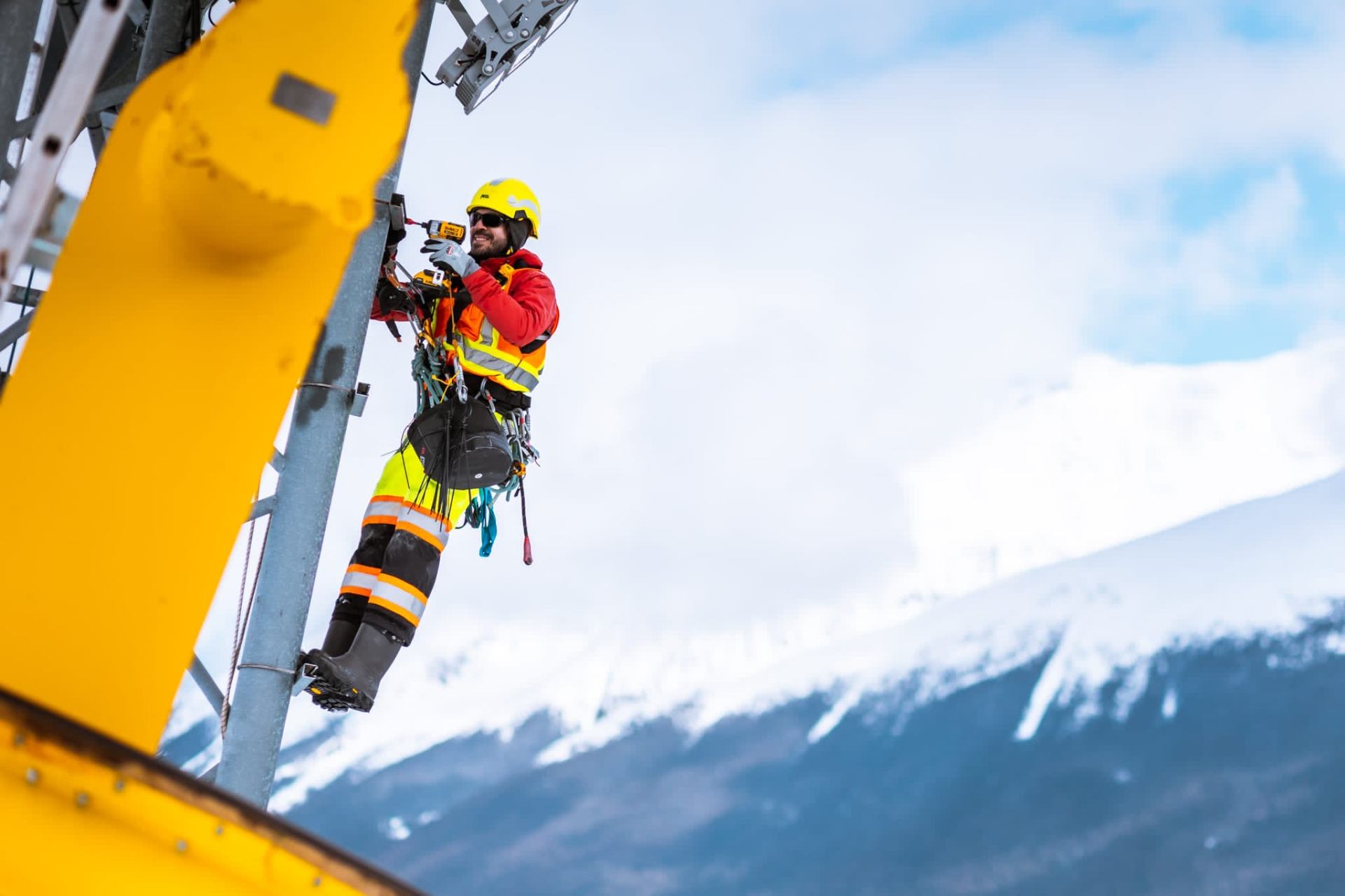 AIM technician working at height on a yellow steel tower with snow-capped mountains behind